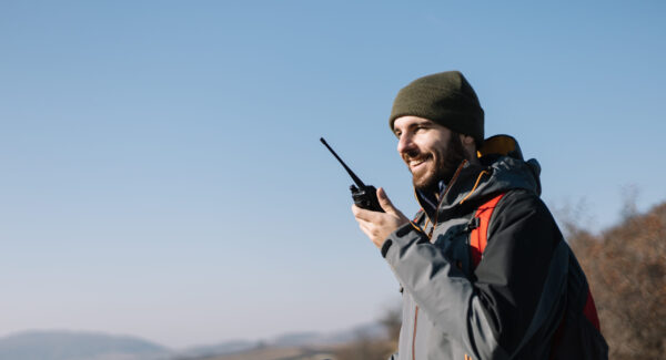 Smiling man speaking on two-way radio for hiking. Young tourist man speaking on mountain walkie-talkie against blue sky.